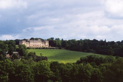 Harewood House is seen on a hill from the south. There is a large grass lawn and woodland in the foreground.