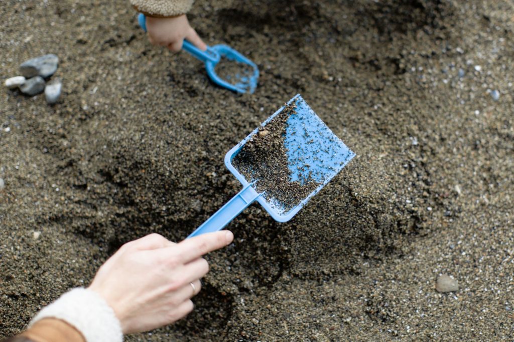 A close up image of an adult and child's hands holding blue digging equipment, digging through sand.