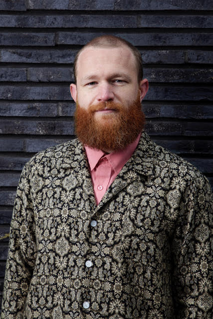a young man with short hair and large ginger beard wearing a patterned grey shirt with a pink collar