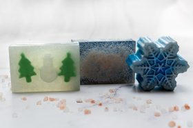 Three different soaps. One soap is clear with two small green christmas trees either side of a white snowman. The middle soap is blue with silver glitter. The last soap is in the shape of snowflake and is blue with silver glitter.