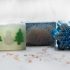 Three different soaps. One soap is clear with two small green christmas trees either side of a white snowman. The middle soap is blue with silver glitter. The last soap is in the shape of snowflake and is blue with silver glitter.