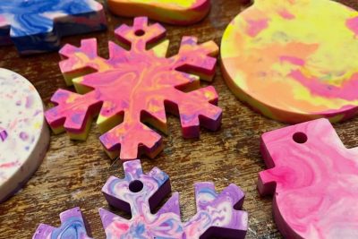 A series of snowflake shaped christmas decorations laid out on a brown background. The snowflakes are pink, yellow and purple.