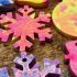 A series of snowflake shaped christmas decorations laid out on a brown background. The snowflakes are pink, yellow and purple.