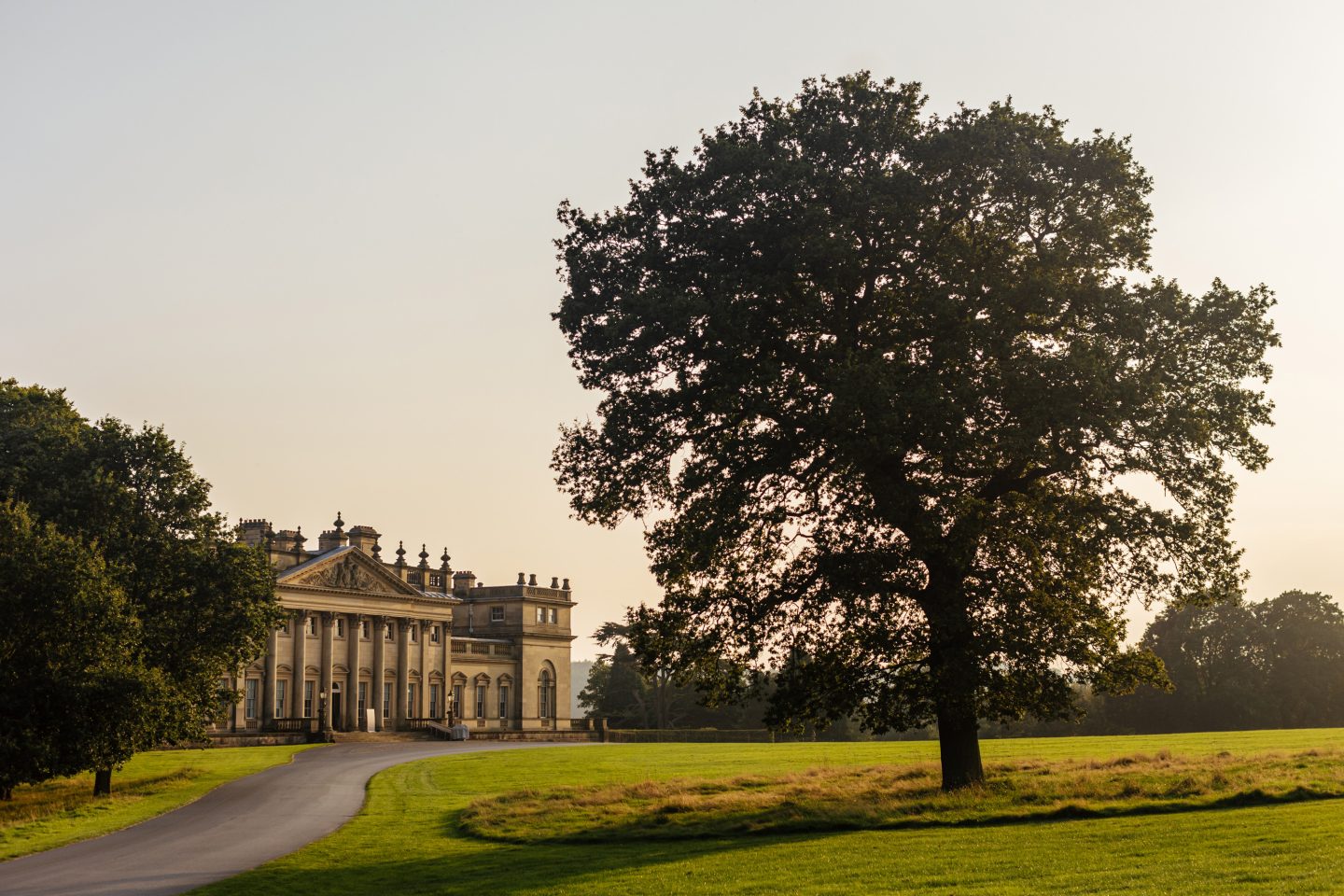 The golden glow of the setting sun bounces off the warm Palladian style country house, casting a dark shadow from a nearby tree.