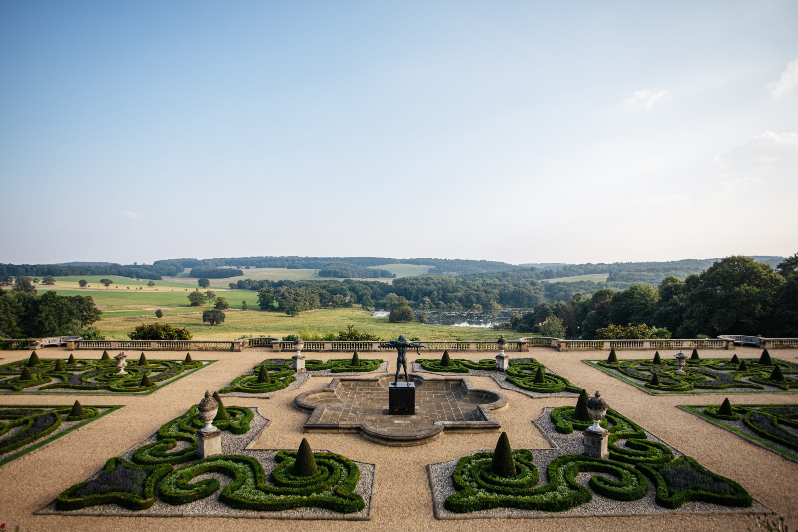 Clear blue skies sit above the rolling Capability Brown landscape with trees dotted across the horizon. Orpheus stands proudly in the middle of an ornate pool of water on the Victorian Terrace.