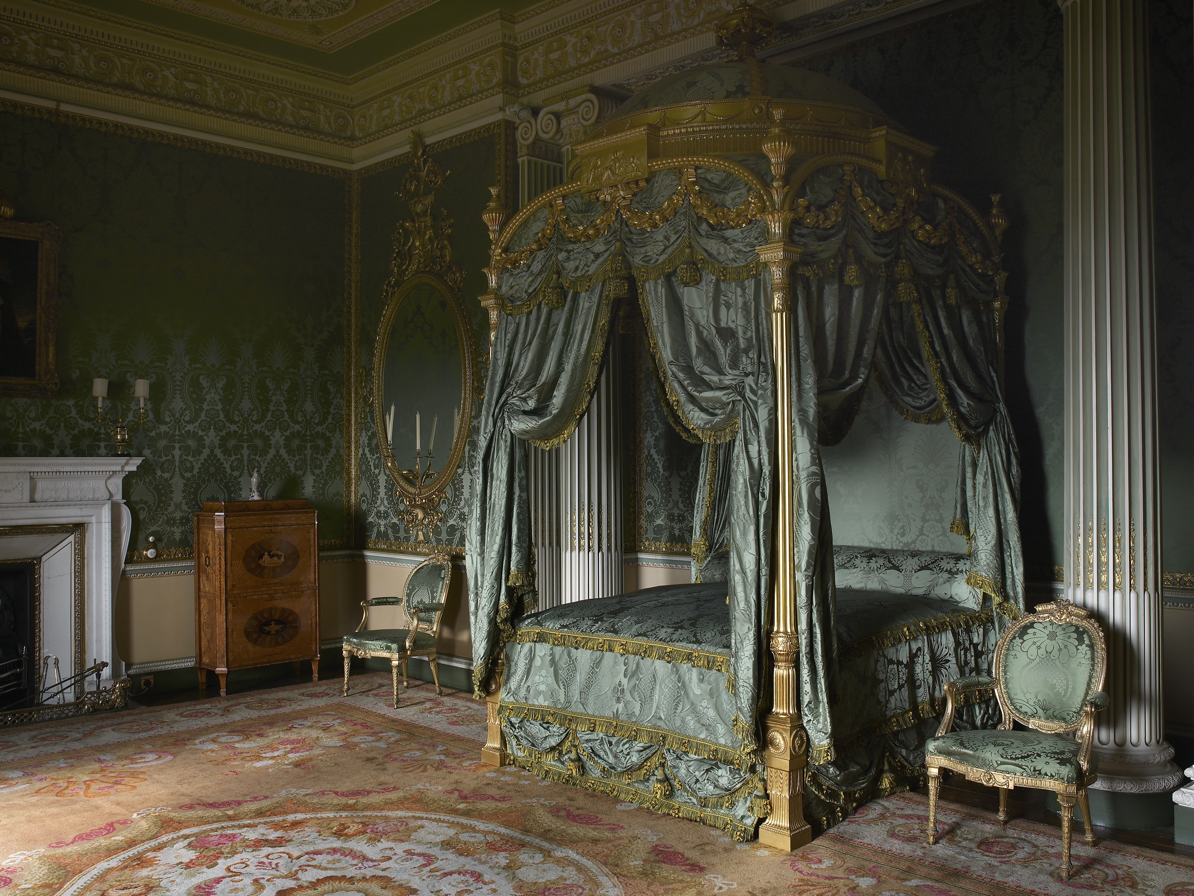 Luxurious bedroom with ornate, green and gold canopy bed, matching chairs, intricate wallpaper, rich decor, wood cabinet, and elegant rug, evoking opulence and grandeur.