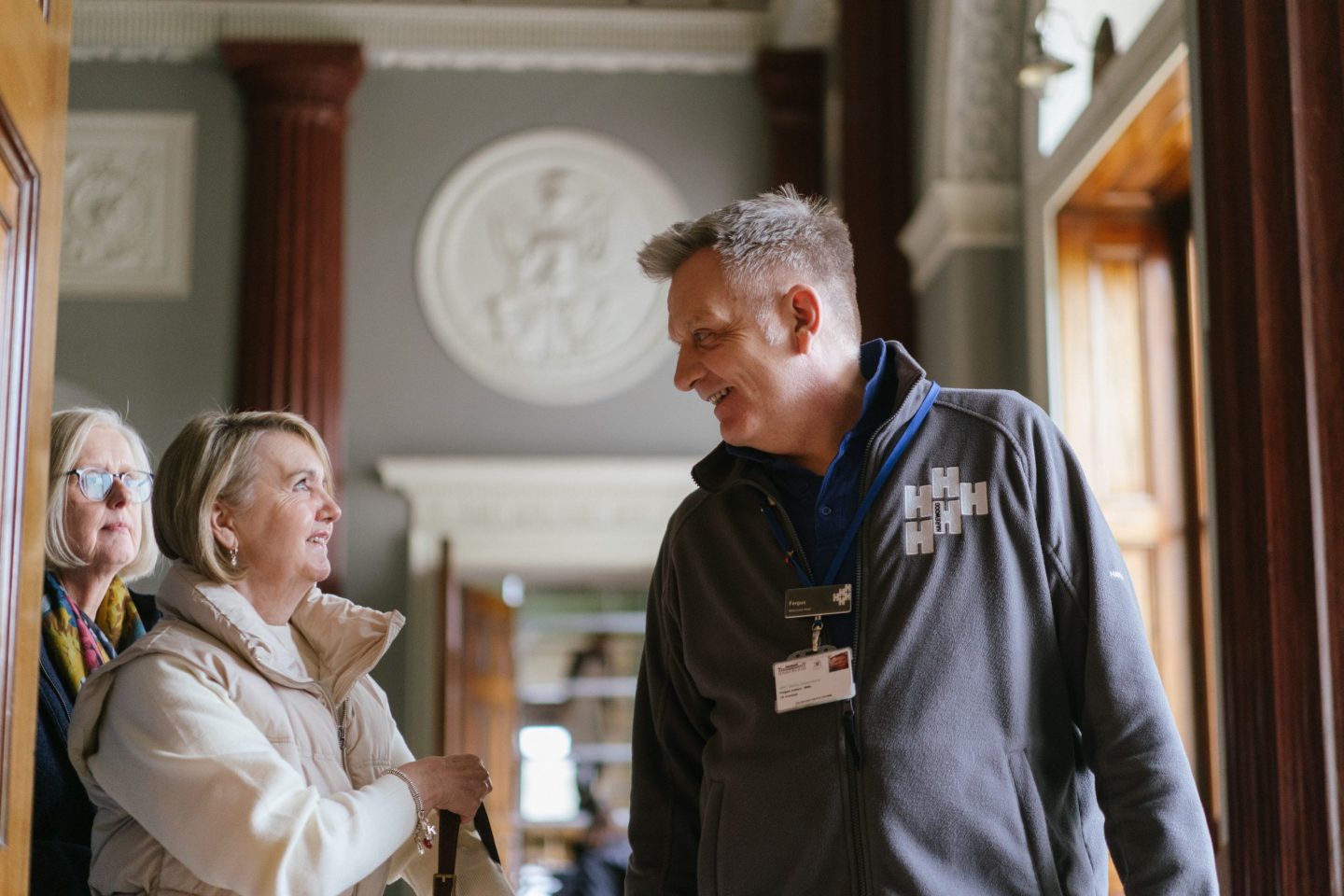A man with short grey hair smiles politely at a women with short blonde hair tucked behind her hear. She wears a cream jumper, cream gilet and pear earrings. The man wears a grey fleece with Harewood logo embroidered on the chest, name badge and key card around his neck.