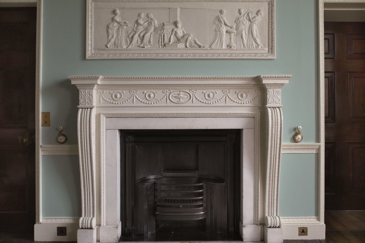 A white, intricately carved mantlepiece below a plasterwork frieze depicting a classical scene.