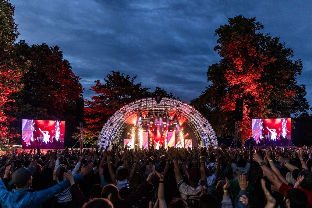 An outdoor concert with a view of the stage with the audience in front with their hands in the air. 2 large screens are either side of the stage with an image of a Queen tribute act. Bahind the stage are trees against a cloudy, night sky