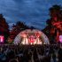 An outdoor concert with a view of the stage with the audience in front with their hands in the air. 2 large screens are either side of the stage with an image of a Queen tribute act. Bahind the stage are trees against a cloudy, night sky