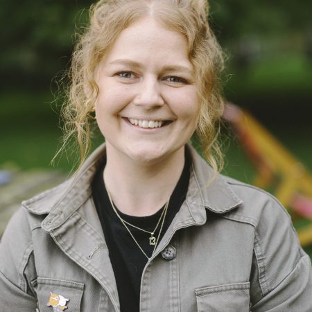 A smiling person with curly blonde hair, wearing a khaki green jacket with a tri colour corgi brooch pinned to the pocket.
