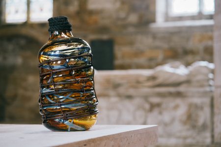 A contemporary glass sculpture in the shape of a large bottle. The glass is a brown syrupy colour with blue streaks swirling around. The bottle is bound by a copper coil that runs around the body, and a coil of rope representing the stopper.