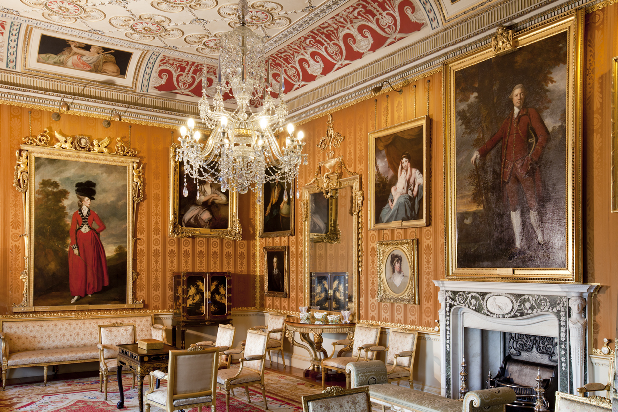 A grand room with cinnamon coloured damask wall hangings, hung with numerous portraits of the Lascelles family. The furniture is upholstered in white.