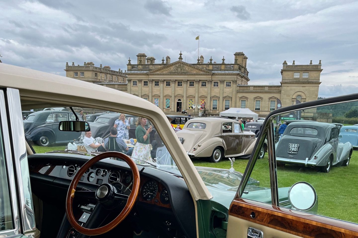 In the front of the picture there is a green Rolls Royce with it's door open, showing the wooden steering wheel and dashboard. Harewood House in the background along with rows of more rolls royce cars.