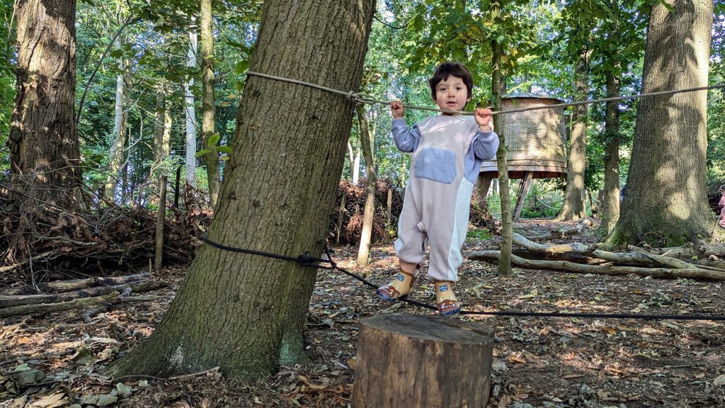 Young child wearing a romper suit stands on a rope bridge between two trees during a Tree Toddlers exploration session.