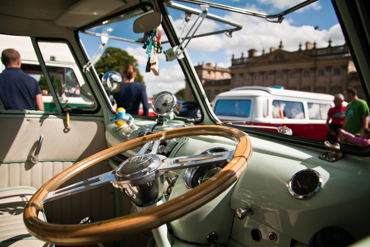 Sage green vintage Volkswagen Beetle parked in front of Harewood House. The dashboard is so clean it's sparkling in the sunshine whilst the wooden steering wheel reflects the golden glow. People wander around in the background, looking at the other vintage Volkswagen cars.