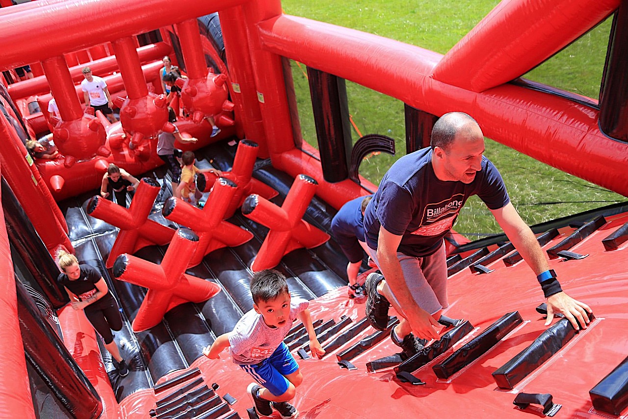 A man and a boy in t-shirts and shorts climb up a bright red and black inflatable obstacle on a sunny day. In the background, participants are making their way past giant inflatable red Xs and inflatable spiky balls suspended from an inflatable bar above the course.